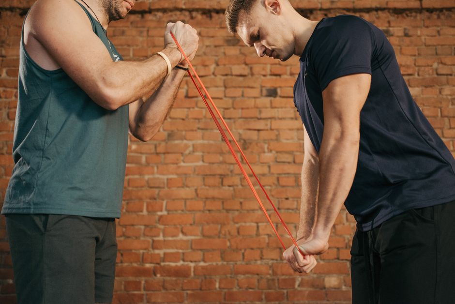 Two men exercising with a resistance band in a brick-walled gym setting.