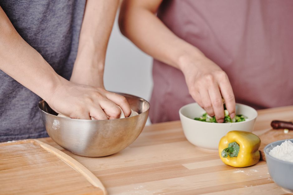 Two people preparing food by kneading dough and cutting vegetables in a kitchen setting.