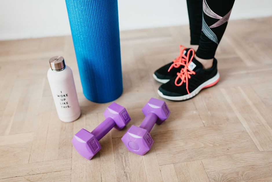 Unrecognizable sportswoman wearing sneakers and black leggings standing with sport mat on wooden floor near dumbbells and water bottle before exercising indoors