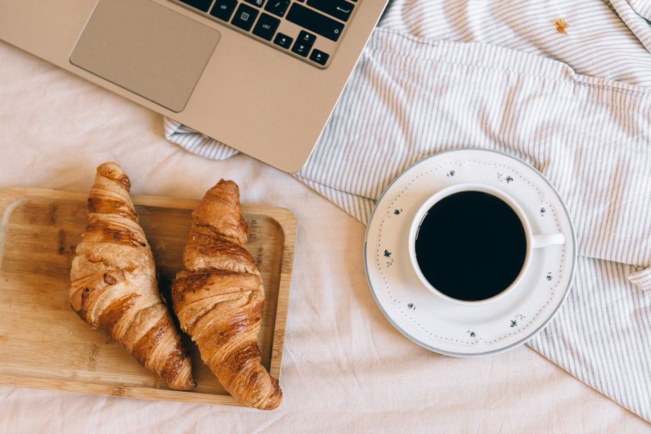 Warm flat lay of croissants with a cup of coffee next to a laptop on a tray.