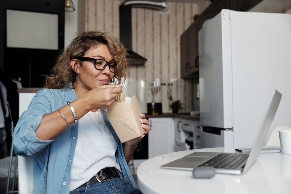 Woman enjoying takeaway pasta at home while working on a laptop. Casual lifestyle setting.