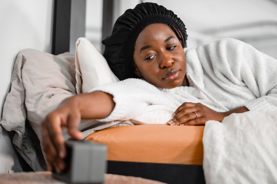 Woman in a cozy robe reaching for alarm clock in bedroom, waking up refreshed.