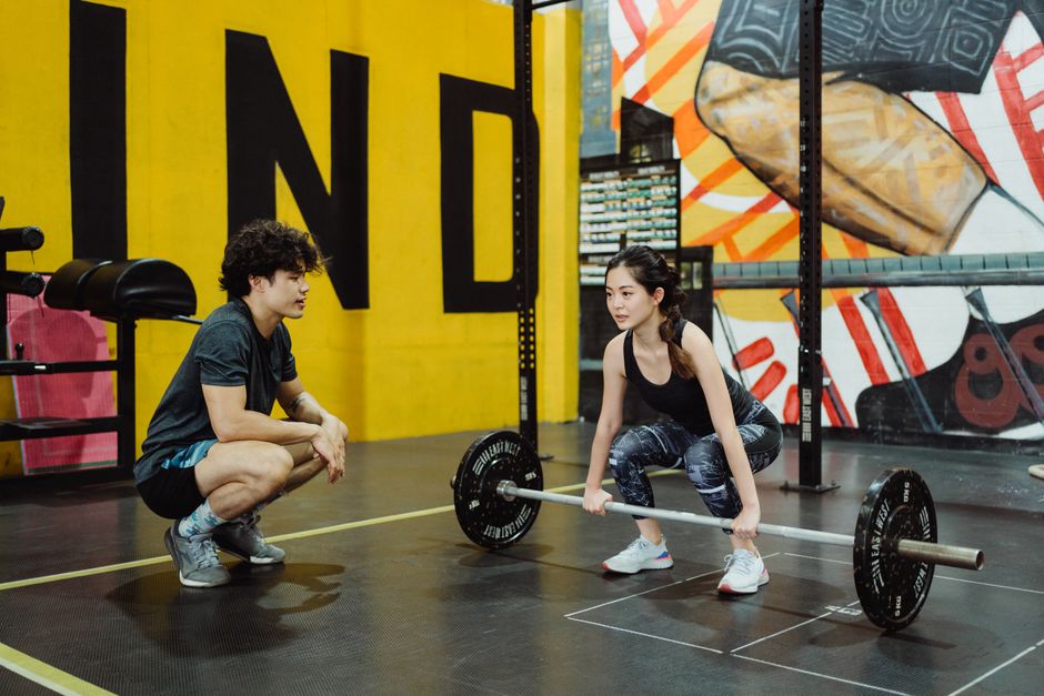 Woman lifting weights with coach's guidance in a vibrant indoor gym setting.