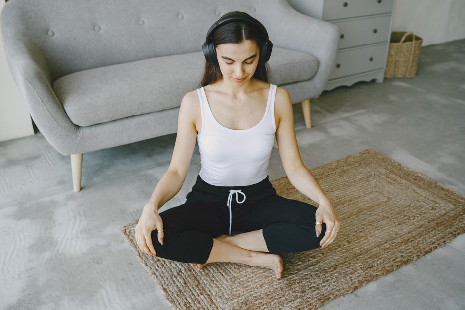 Woman meditating indoors on a rug with headphones, promoting relaxation and wellness.