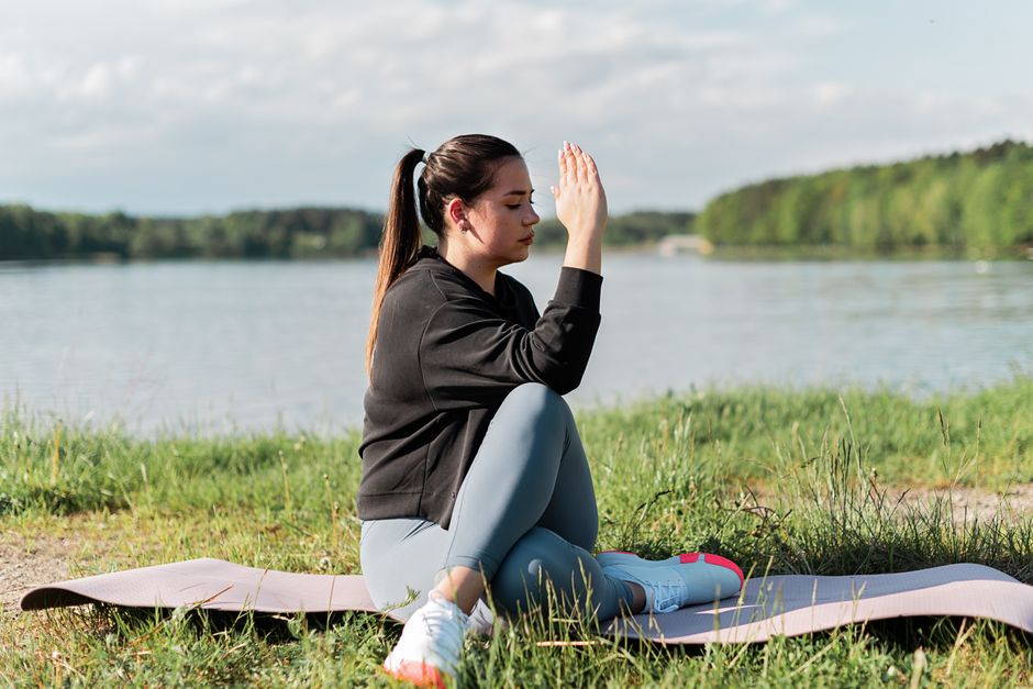Young woman meditating outdoors on a yoga mat by the lake, enjoying a peaceful moment.