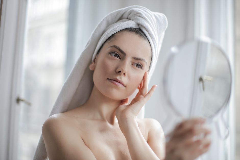 Young woman with a towel on her head looking into a handheld mirror indoors, promoting self-care.