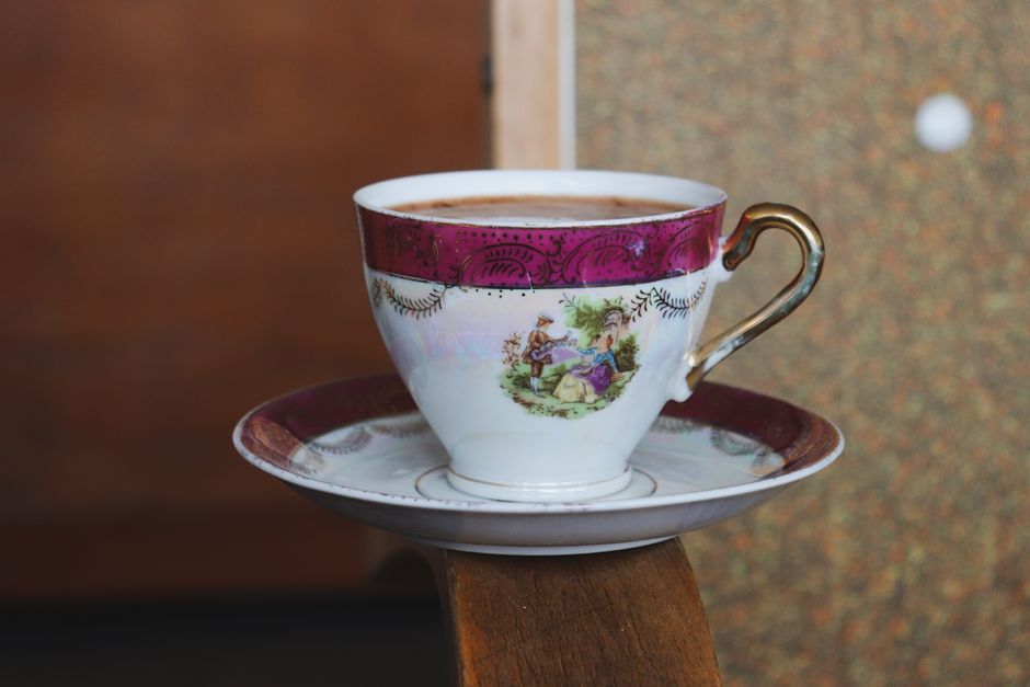 A beautifully decorated porcelain teacup filled with coffee, resting on a saucer, in a cozy indoor setting.
