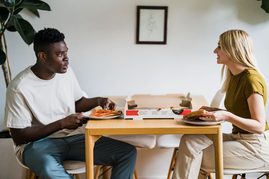 A cheerful interracial couple shares a relaxing pizza meal at home, sitting across a wooden table.