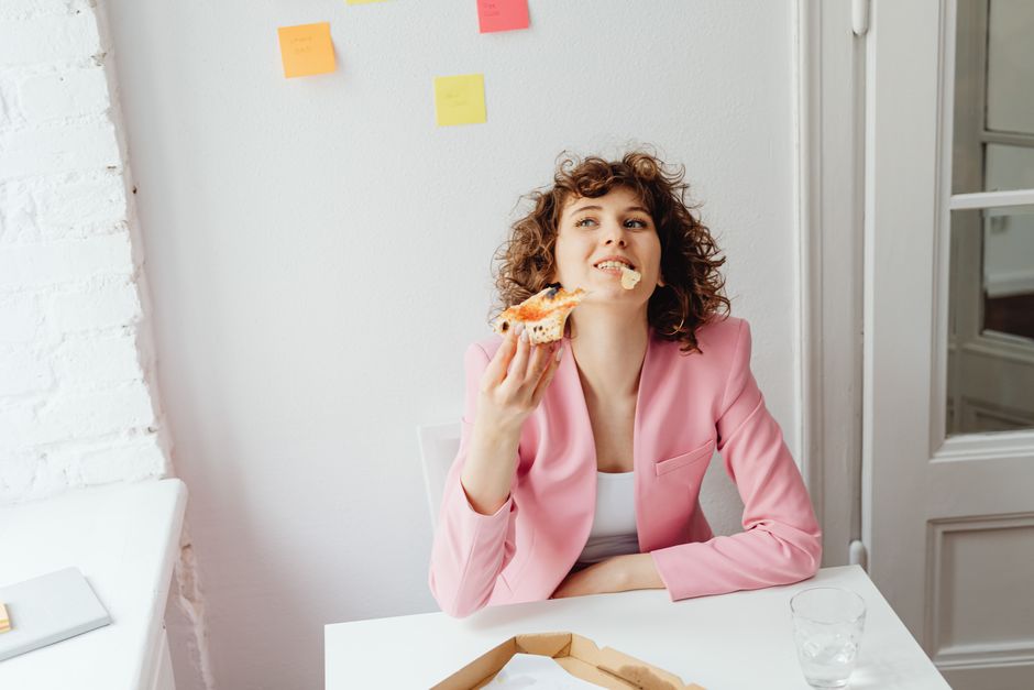 A cheerful woman in a pink blazer takes a moment to enjoy pizza while working from home.