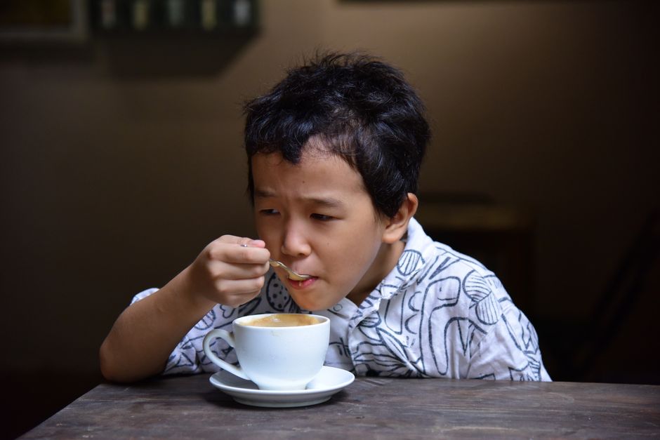 A child carefully sipping coffee from a cup in a cozy indoor setting.