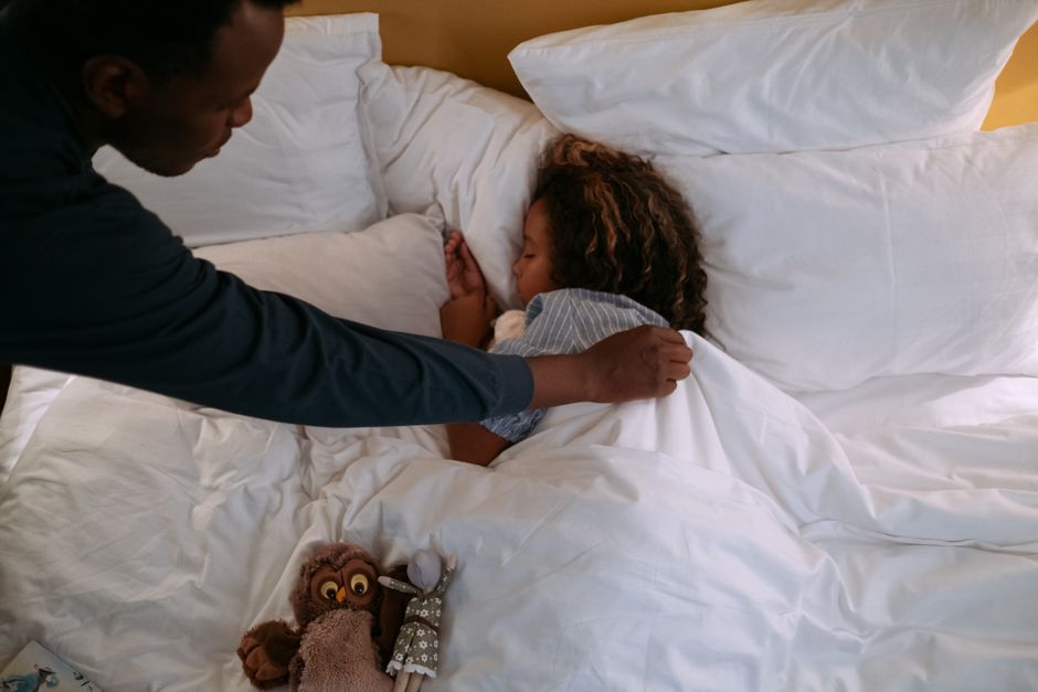 A father lovingly tucks his sleeping daughter into bed, ensuring her comfort.