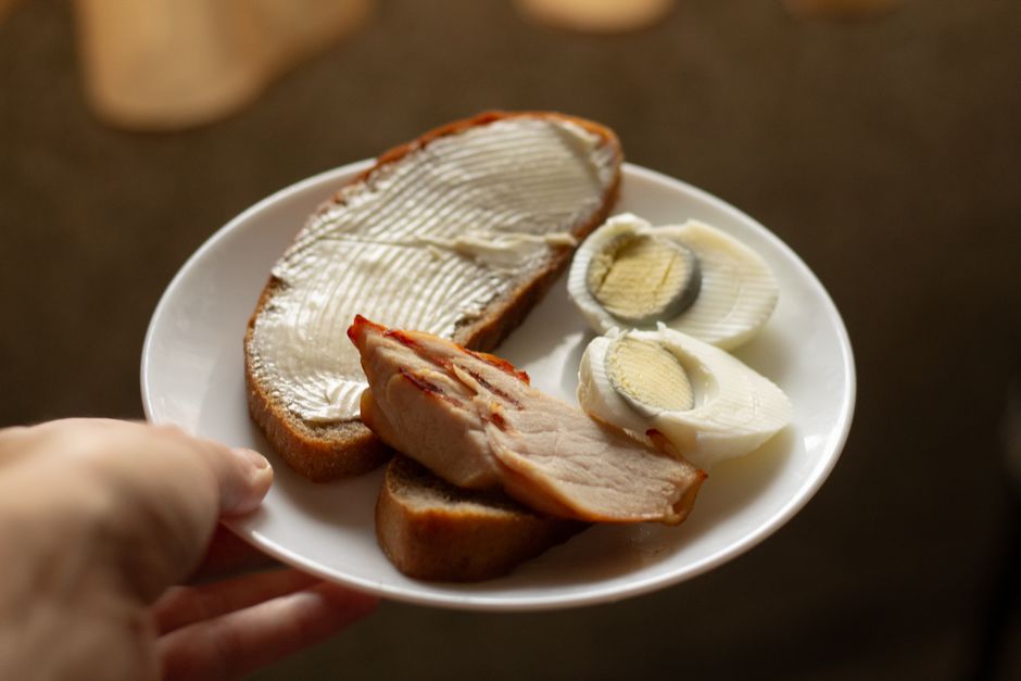 A hand holds a plate featuring buttered bread, sliced egg, and a meat piece.