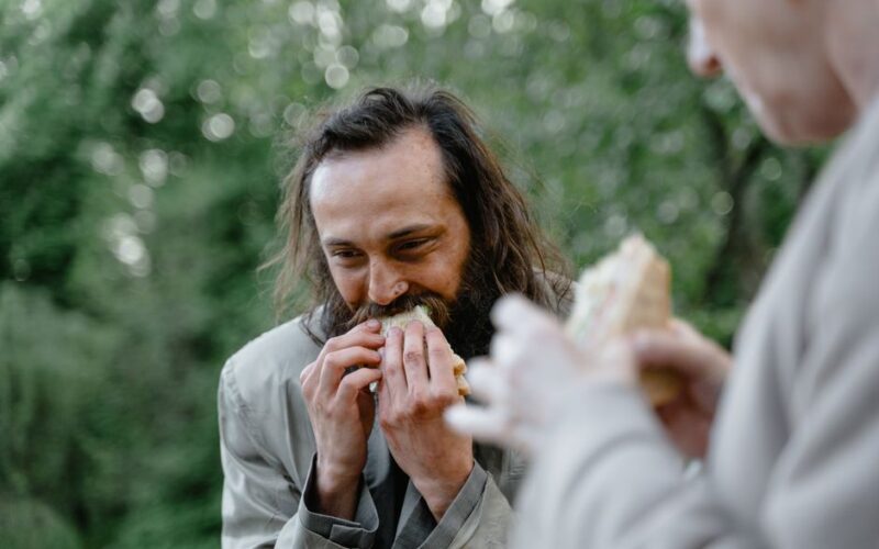 A man happily eats a sandwich outside in a green and natural setting.