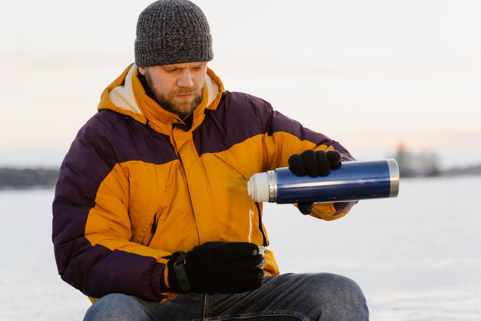 A man in winter clothing pours hot drink from a thermos in snowy landscape.