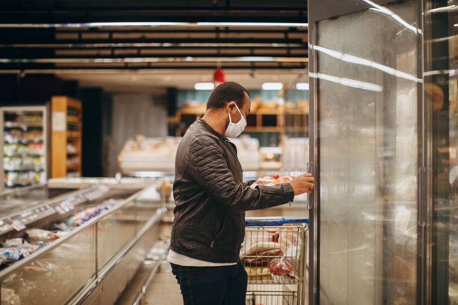 A man wearing a mask shops in a supermarket, selecting items from a refrigerated section.