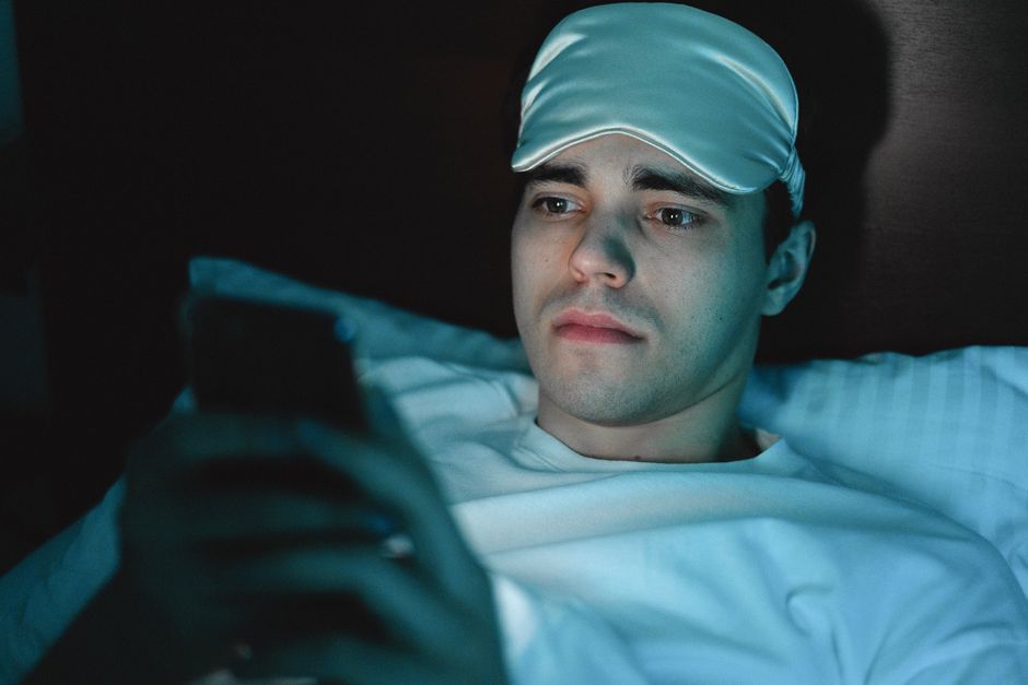 A man wearing a sleeping mask uses his smartphone while lying in bed at night.