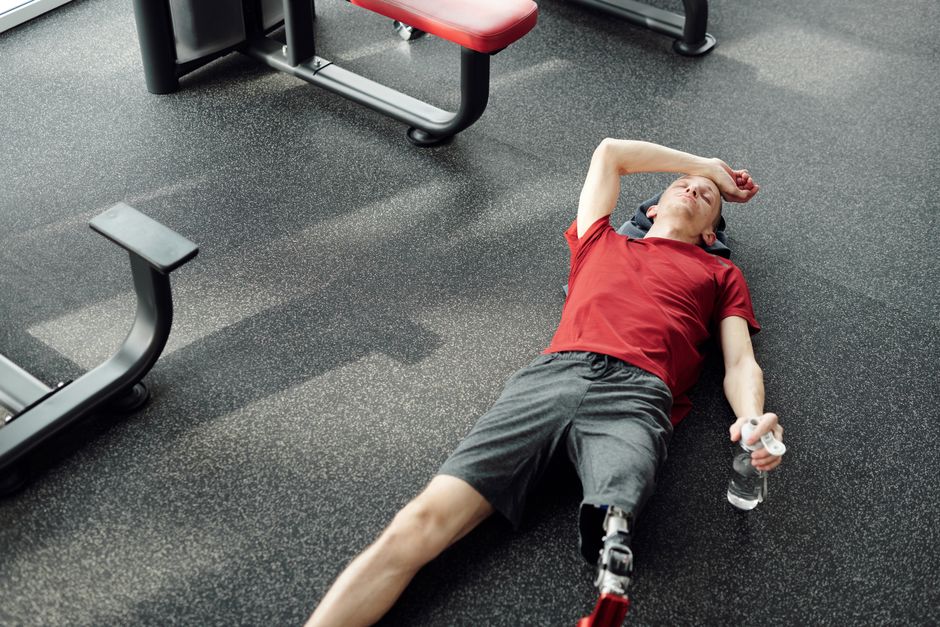 A man with a prosthetic leg rests on the gym floor, holding a water bottle after an intense workout.