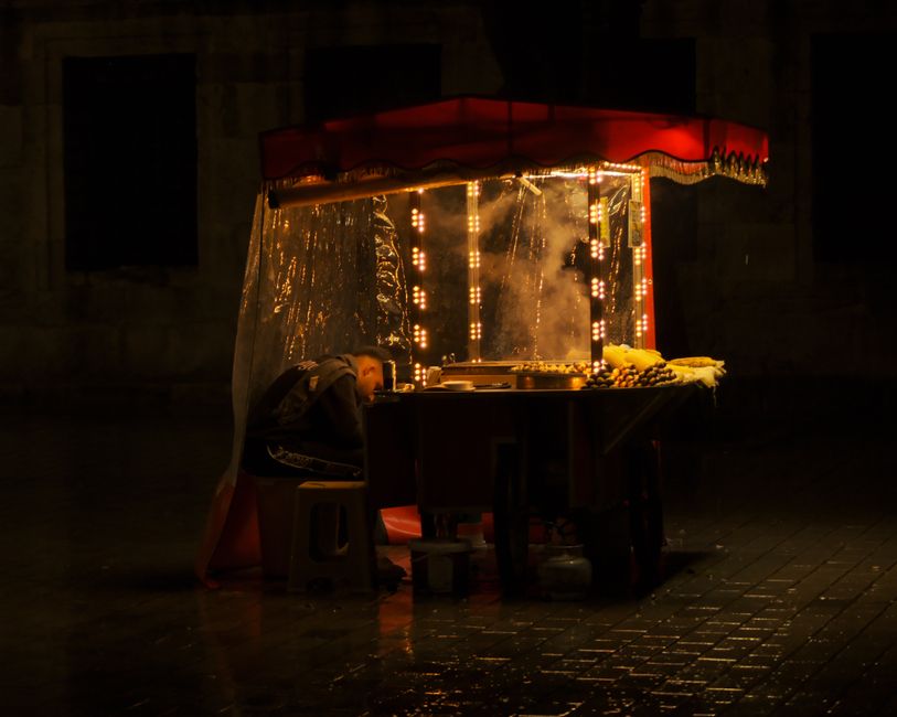 A market vendor sleeping at a street food stall under warm lights at night.