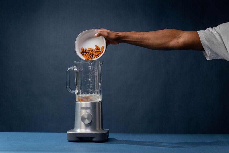 A person adds almonds to a blender, preparing ingredients for a healthy drink in a minimalist studio setting.