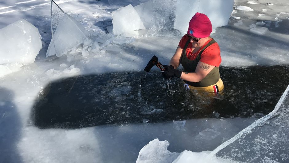 A person dressed in winter gear cutting through thick ice on a frozen lake.