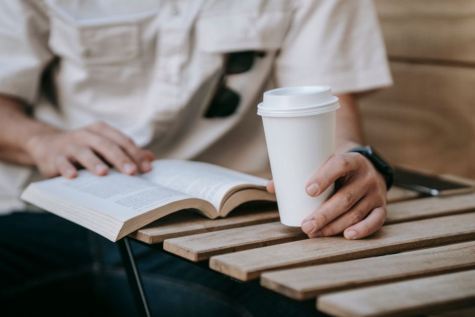 A person enjoys coffee while reading a book outdoors on a wooden table.