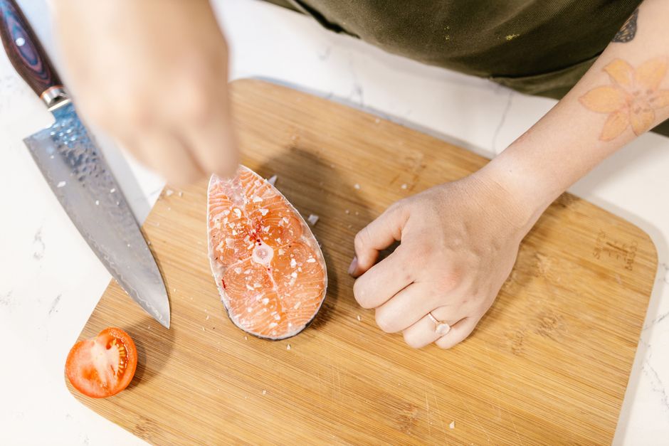 A person preparing salmon steak on a wooden board with a knife and tomato slice.