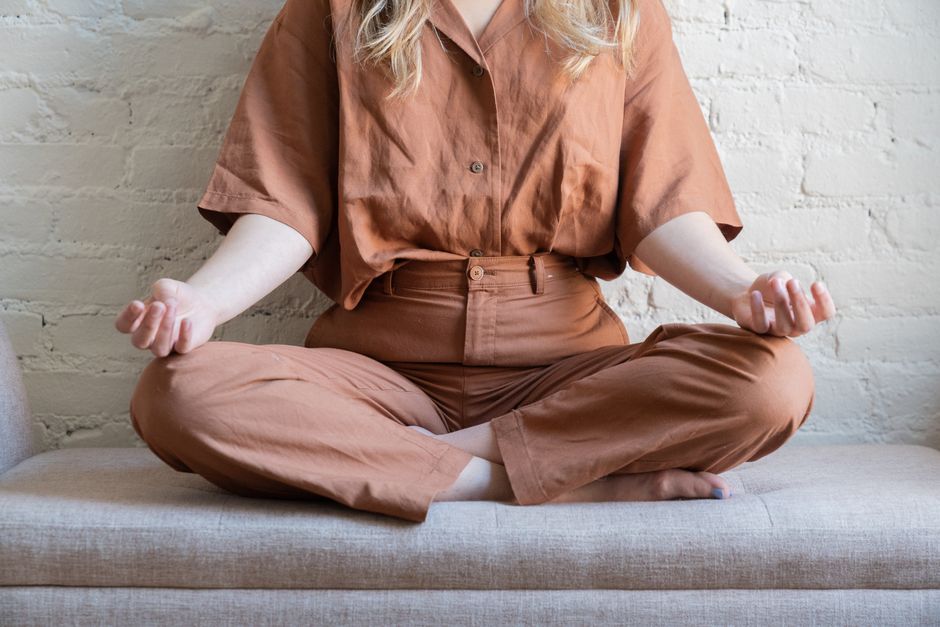 A person sits meditatively, hands in mudra, dressed in warm earth tones, indoors.