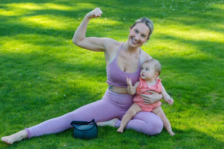 A smiling mother flexes playfully while sitting with her baby on a sunny lawn.