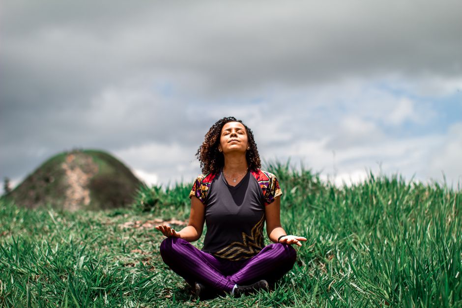 A woman enjoys peaceful meditation in a grassy field under a cloudy sky in Brazil.