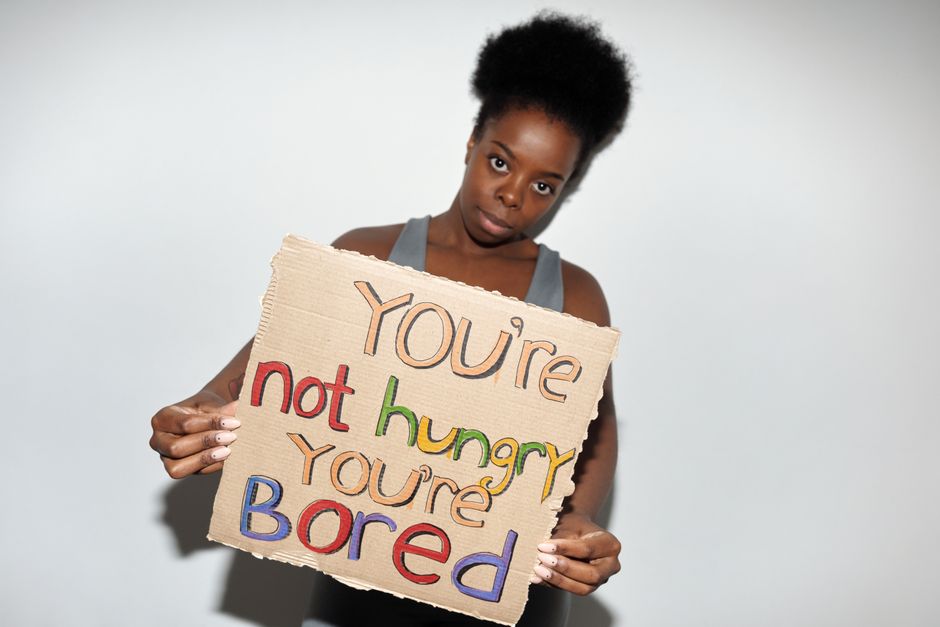 A woman holds a cardboard sign with the message 'You're not hungry, you're bored' in a studio setting.
