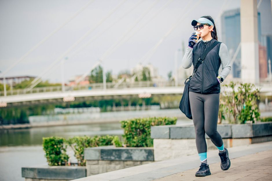 A woman in athletic wear enjoys a drink while jogging along a riverside path.