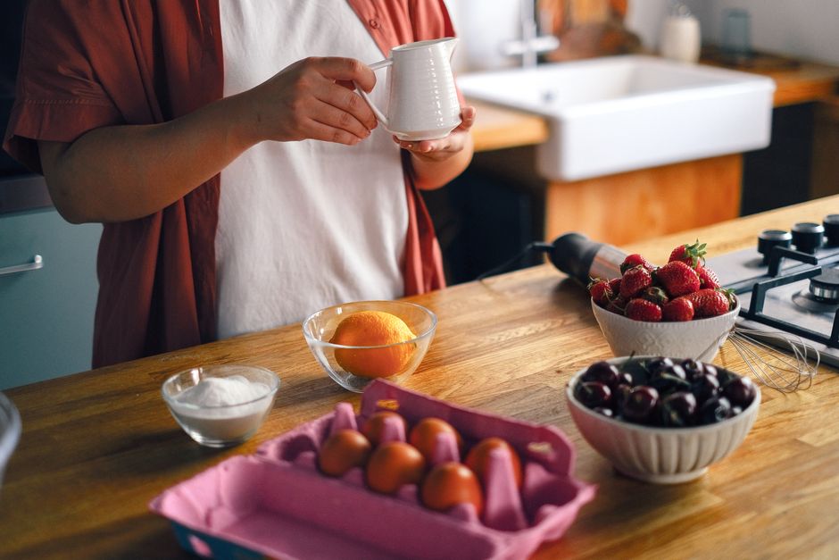 A woman in the kitchen holds a cup surrounded by fresh ingredients like eggs and fruit.