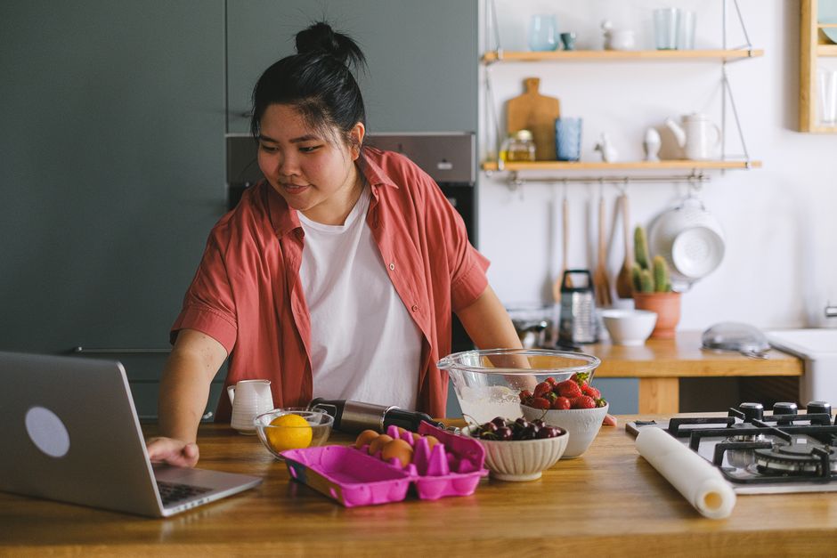 A young adult using a laptop while preparing food in a modern kitchen setting.