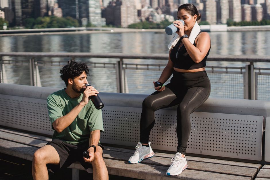 A young couple takes a break by the waterfront to hydrate after exercising outdoors in a sunny city setting.