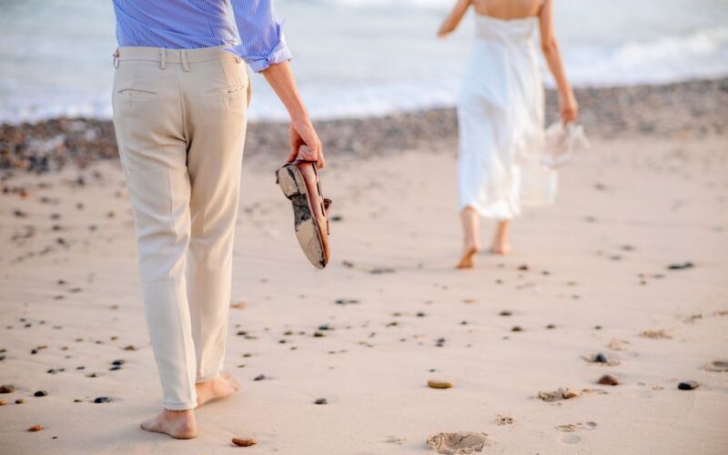 A young couple walks barefoot on a sandy beach, holding shoes in hand, enjoying a serene day by the ocean.