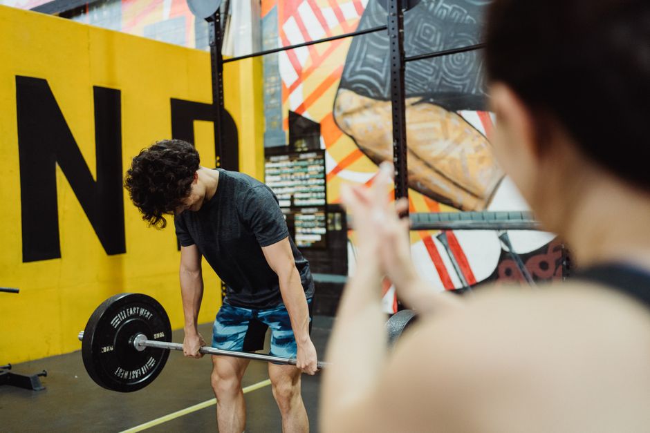 A young man performs a deadlift in a vibrant gym, watched by a supporter.