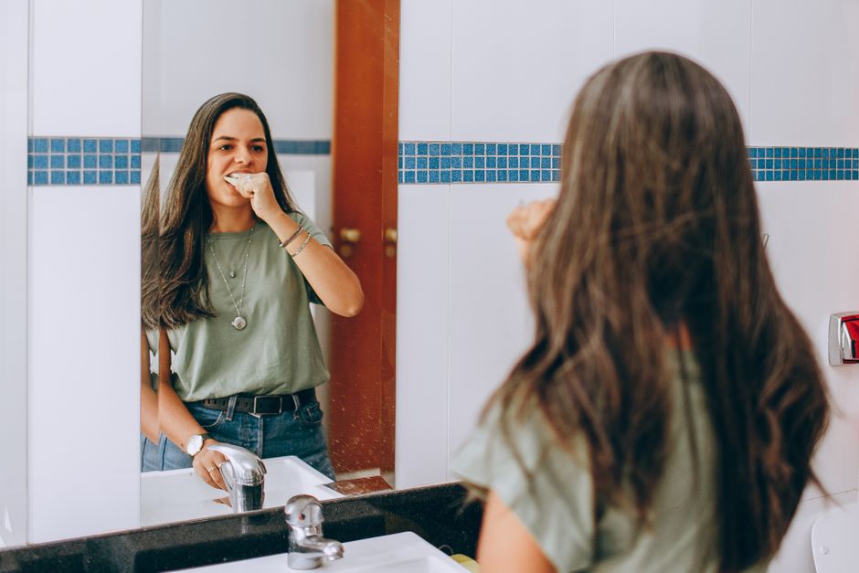 A young woman brushes her teeth, looking in the bathroom mirror. Self-care and hygiene concept.