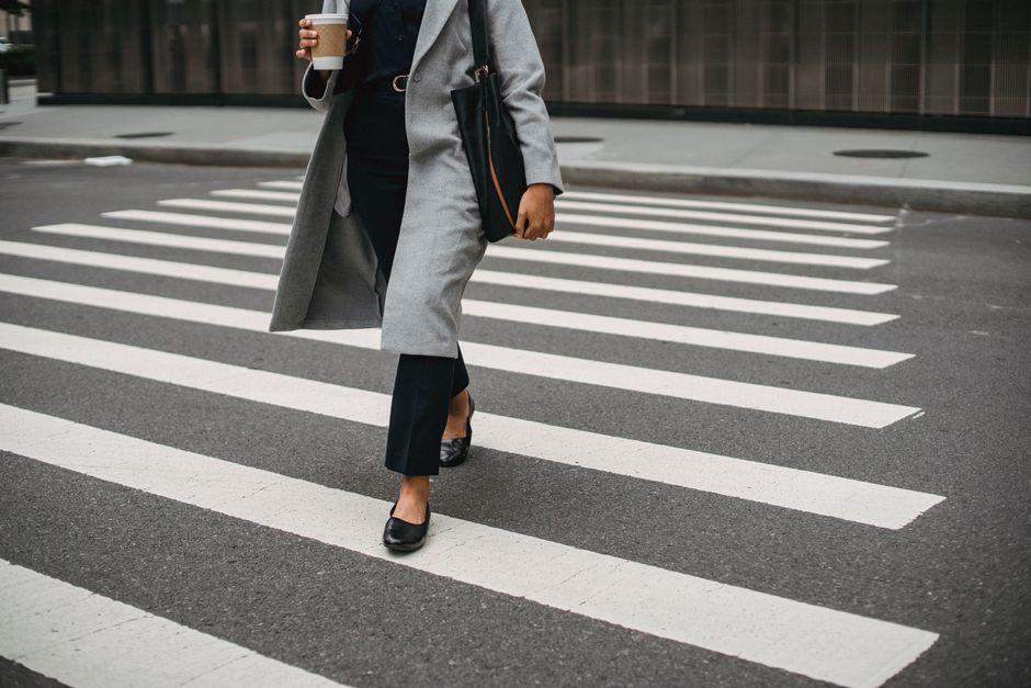 Adult in gray coat crossing city street with coffee and tablet in hand.