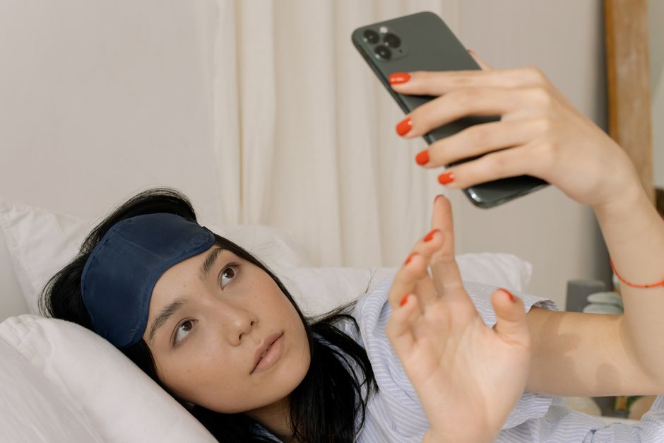An Asian woman with a sleep mask relaxing in bed, using a smartphone indoors.