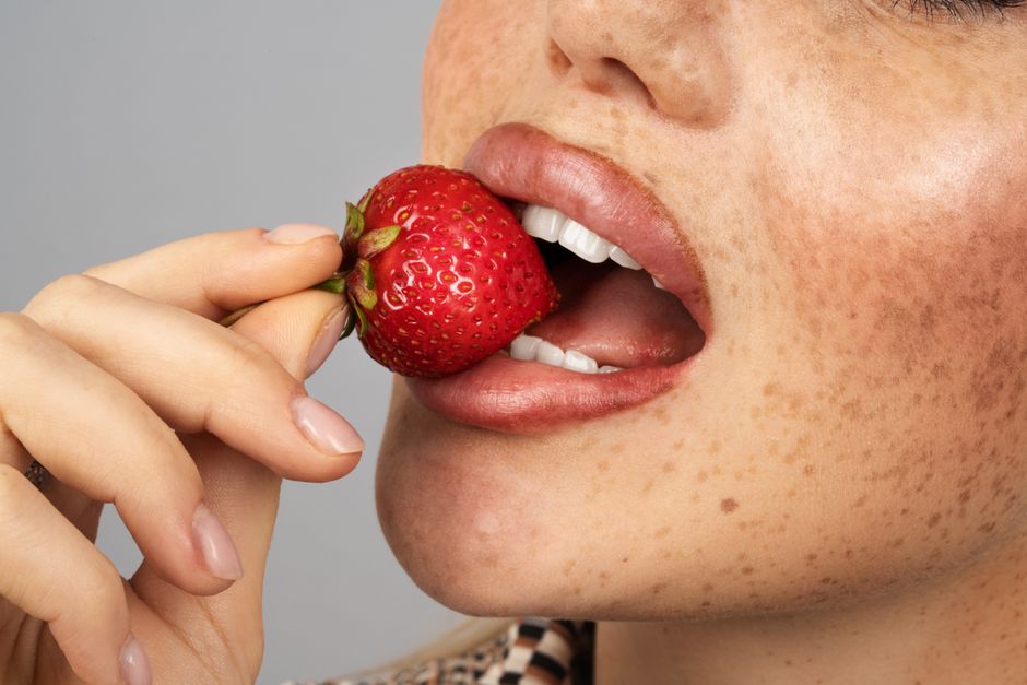 An intimate close-up of a woman enjoying a ripe strawberry with freckles visible on her face.