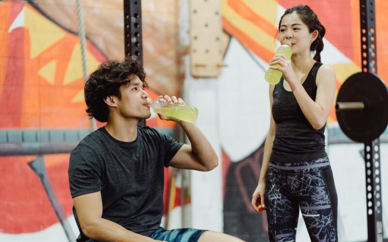 Asian couple in gym attire drinking energy drinks after workout. Vibrant and lively setting.