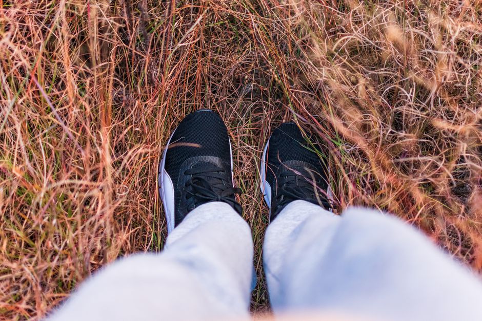 Black sneakers on a person standing amidst tall grass, showcasing casual fashion vibe.