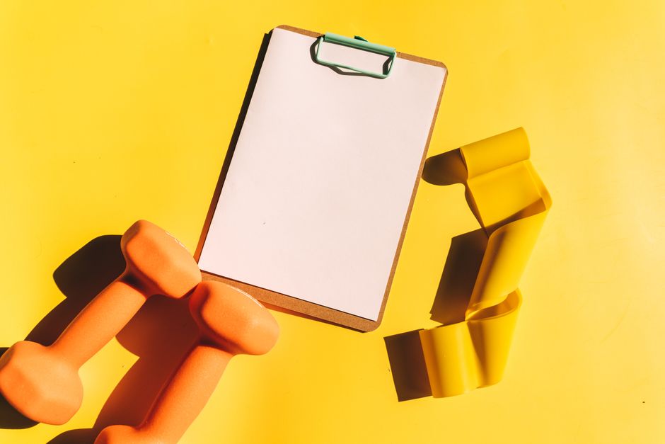 Bright flat lay of orange dumbbells, clipboard, and yellow resistance band on yellow background.