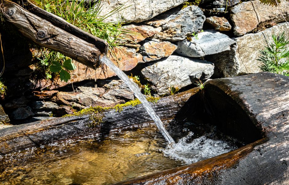 Clear water flows from a rustic wooden spout into a basin, set against a stone wall.