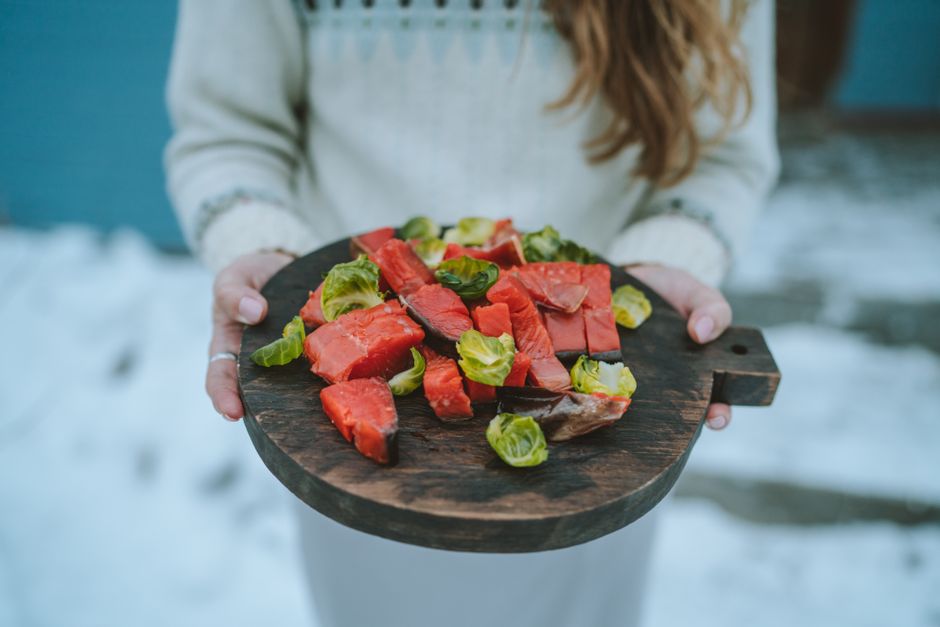 Close-up of a person holding a wooden board with fresh salmon and Brussels sprouts on a snowy day.