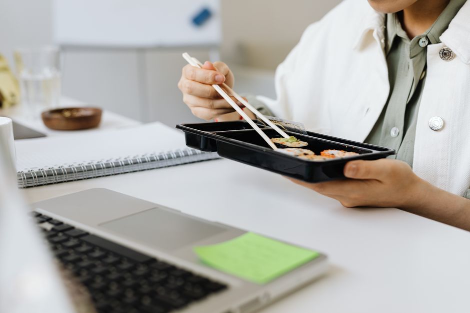 Close-up of a person using chopsticks to eat sushi at a desk with a laptop.