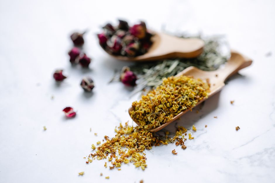 Close-up of dried herbs and flowers in wooden spoons on a marble surface.