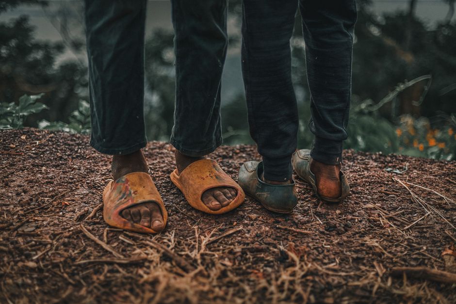 Close-up shot of two kids wearing worn sandals standing on earthy ground outdoors.