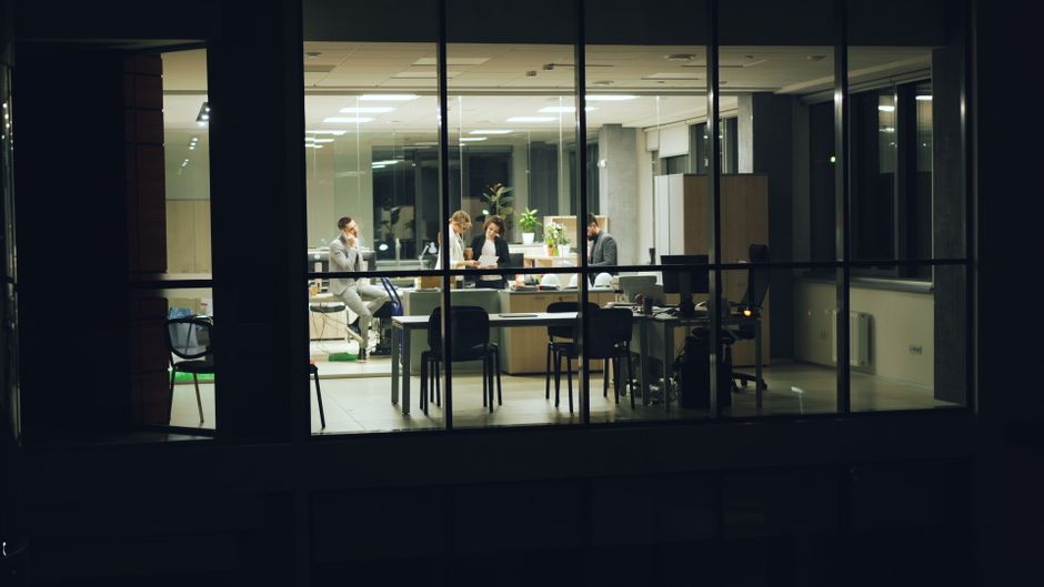 Colleagues collaborate in a glass-walled office at night, focused on tasks.