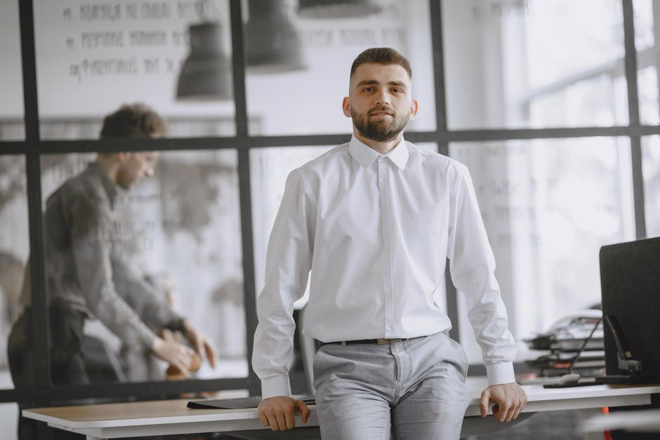 Confident man in white shirt leaning on desk in modern office with glass partitions.